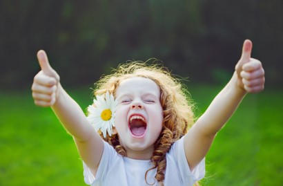 Little girl outside with a daisy in her hair, arms wide open with two thumbs up, smiling in appreciation and joy