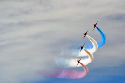 Four jets flying in synchronization, each with a different color contrail: blue, white, white, red/pink