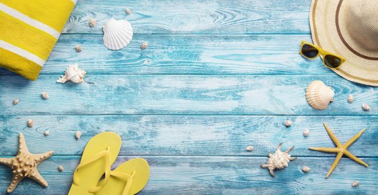 Yellow flip-flops on a pastel torquoise boardwalk with a yellow beach towel, yellow sunglasses, straw beach hat, and various seashells sprinkled about